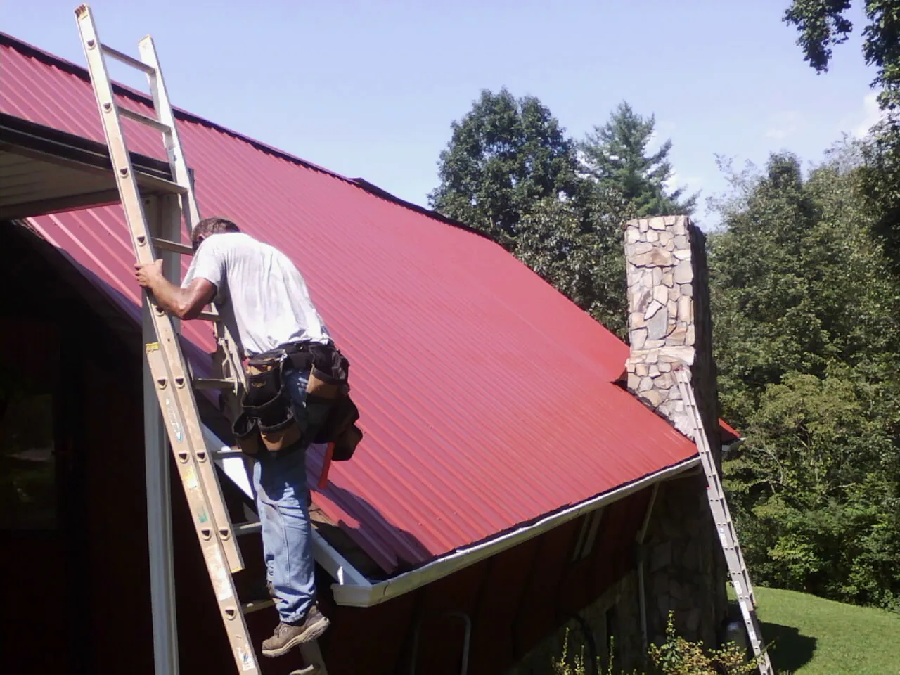 Roofer on a ladder beside a red metal roof and stone chimney — Soffit & Fascia Repair by Roofing Brothers in Lake Lynn